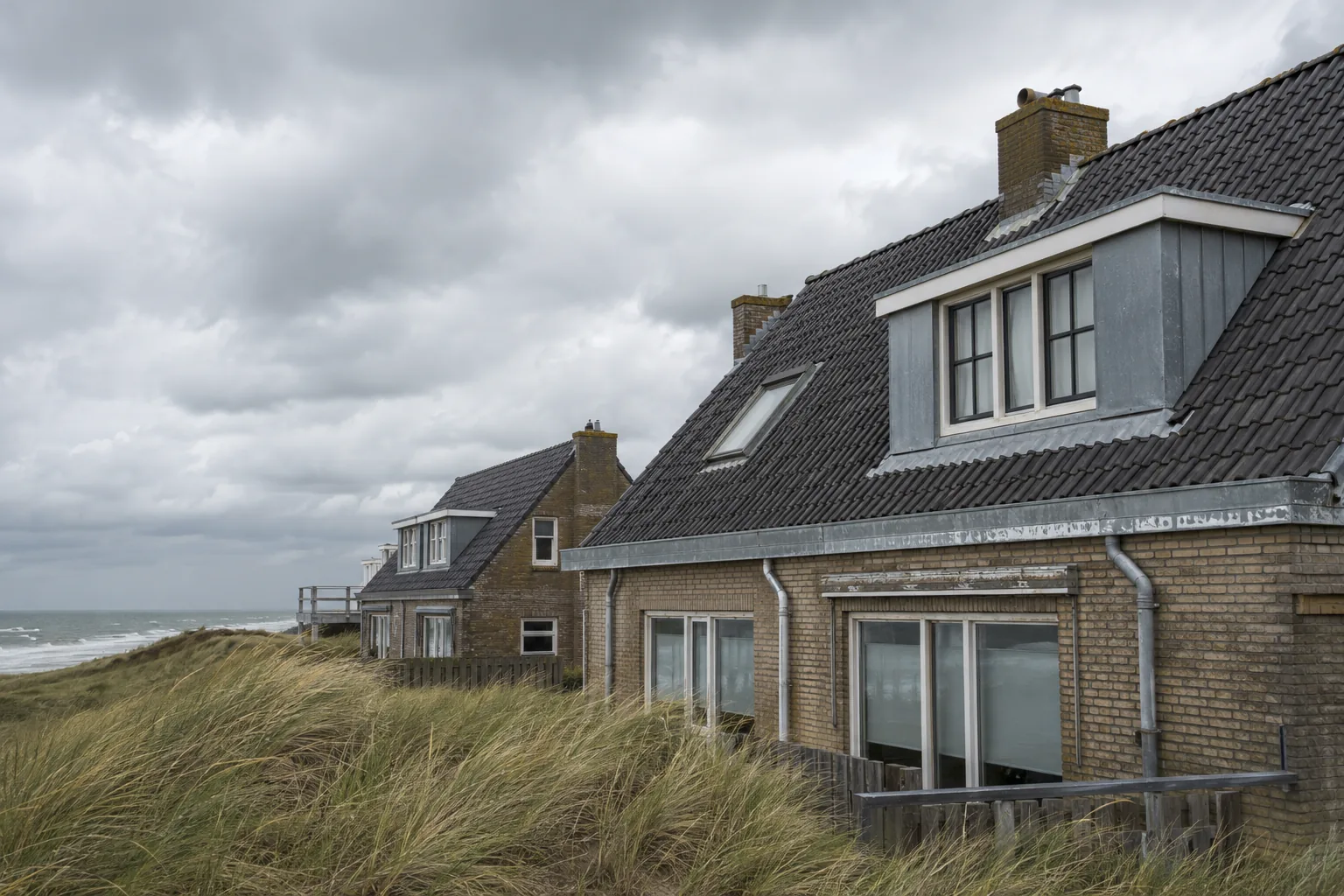 Nederlandse kustwoningen met pannendaken en zinken goten achter de duinen, met helmgras in de wind onder een bewolkte hemel
